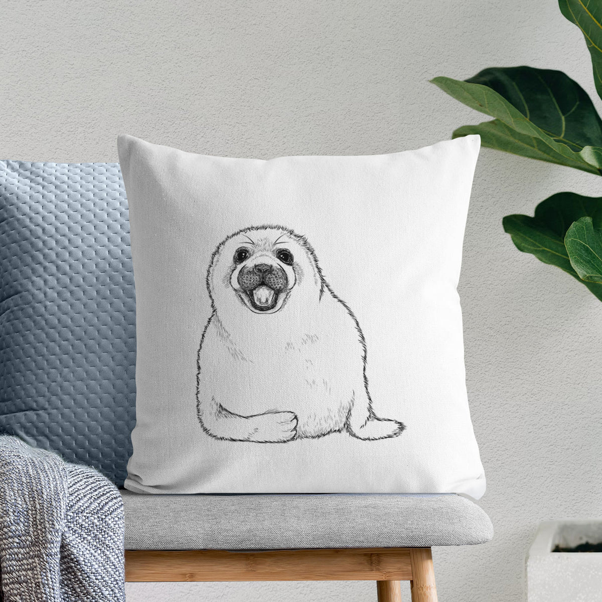 White pillow cover printed with a black ink illustration of a smiling harp seal pup lying down, on a bench beside plants.