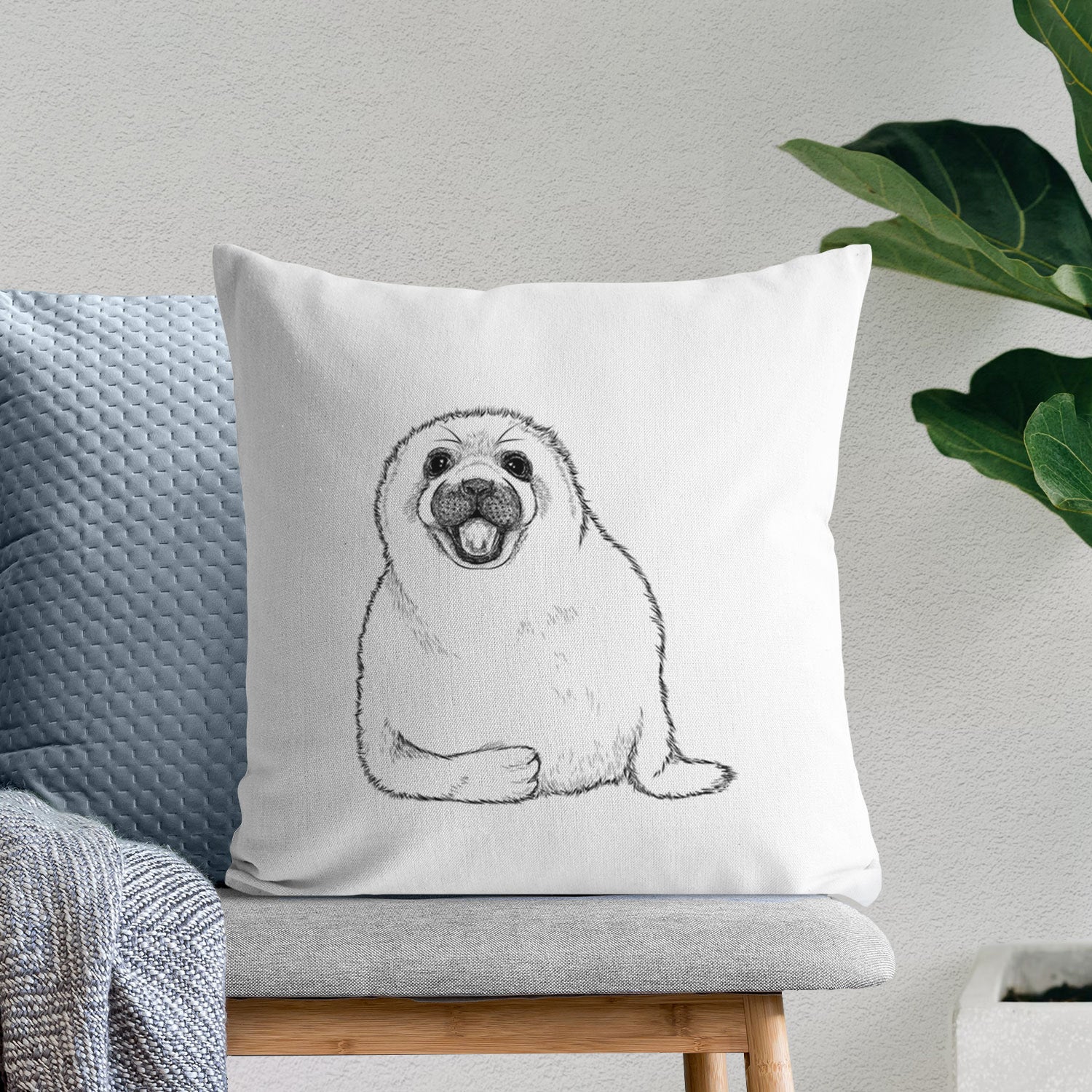 White pillow cover printed with a black ink illustration of a smiling harp seal pup lying down, on a bench beside plants.