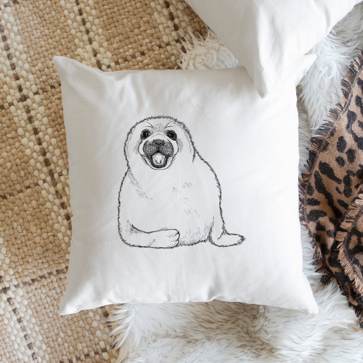 White pillow cover printed with a black ink illustration of a smiling harp seal pup lying down, on a woven chair.