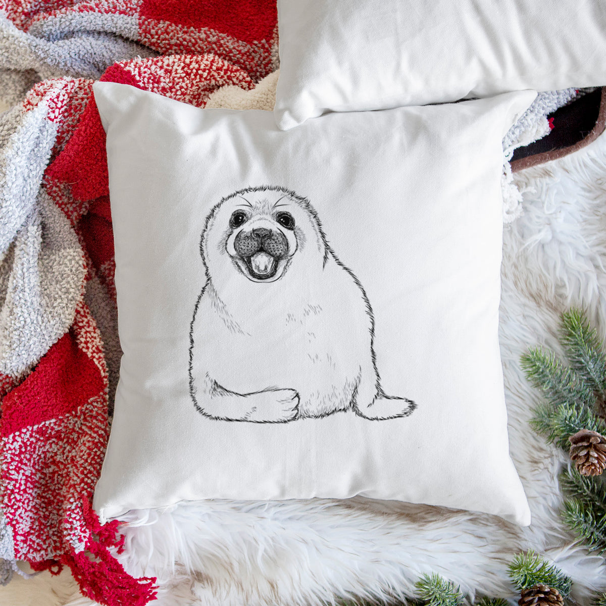 White pillow cover printed with a black ink illustration of a smiling harp seal pup lying down, on a woven chair.