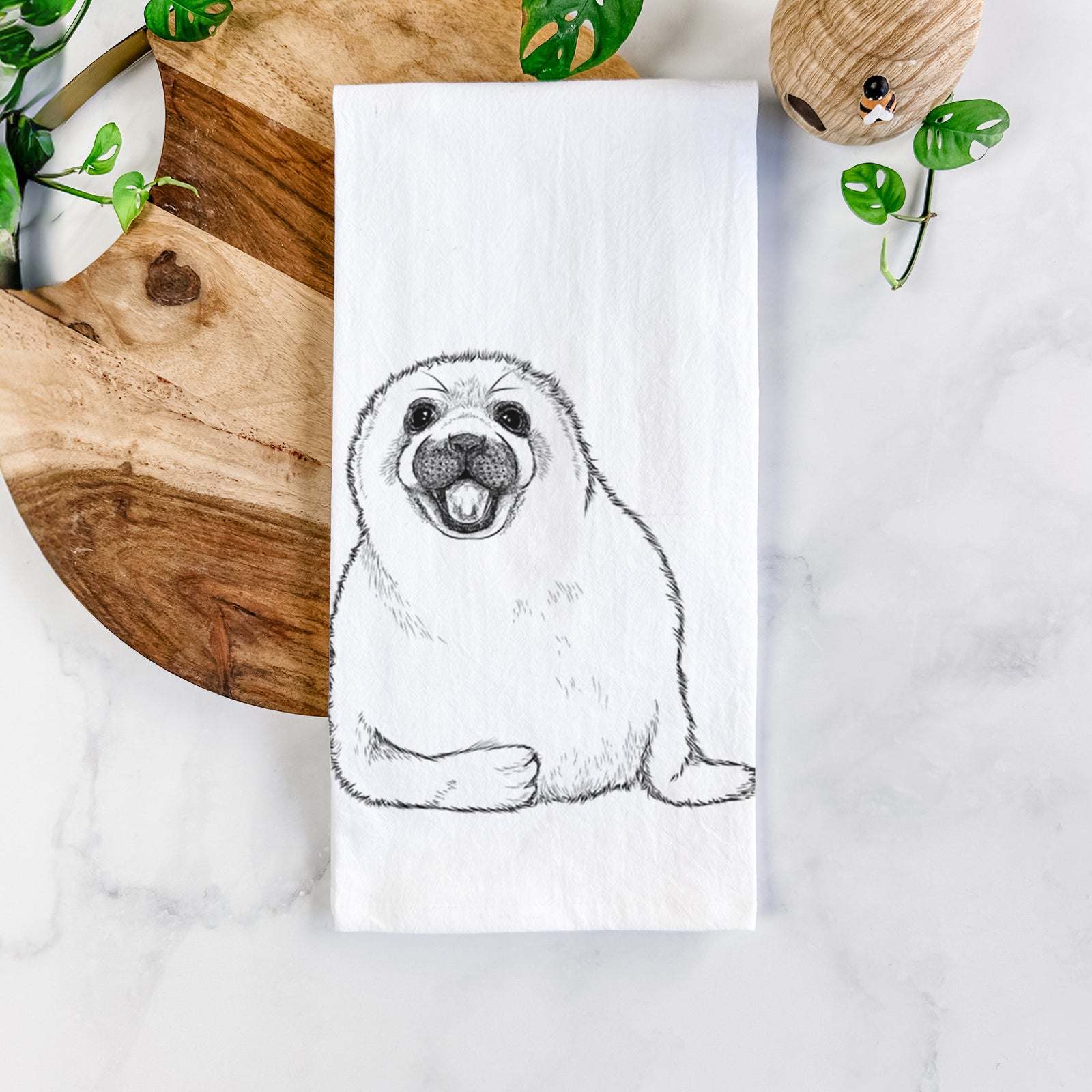 White cotton tea towel printed with a black ink illustration of a smiling harp seal pup lying down, on a marble counter beside a round wooden cutting board and greenery.
