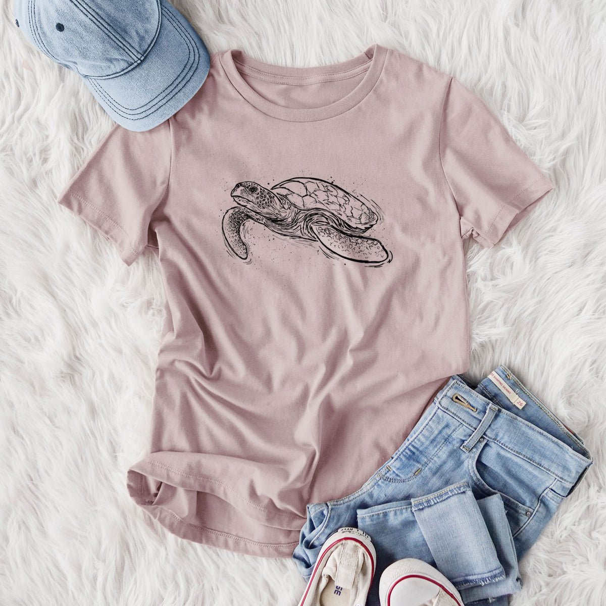 Women's relaxed fit cotton t-shirt printed with a black ink illustration of a hawksbill sea turtle swimming, with a patterned shell, styled flat on a white faux fur rug with jeans.