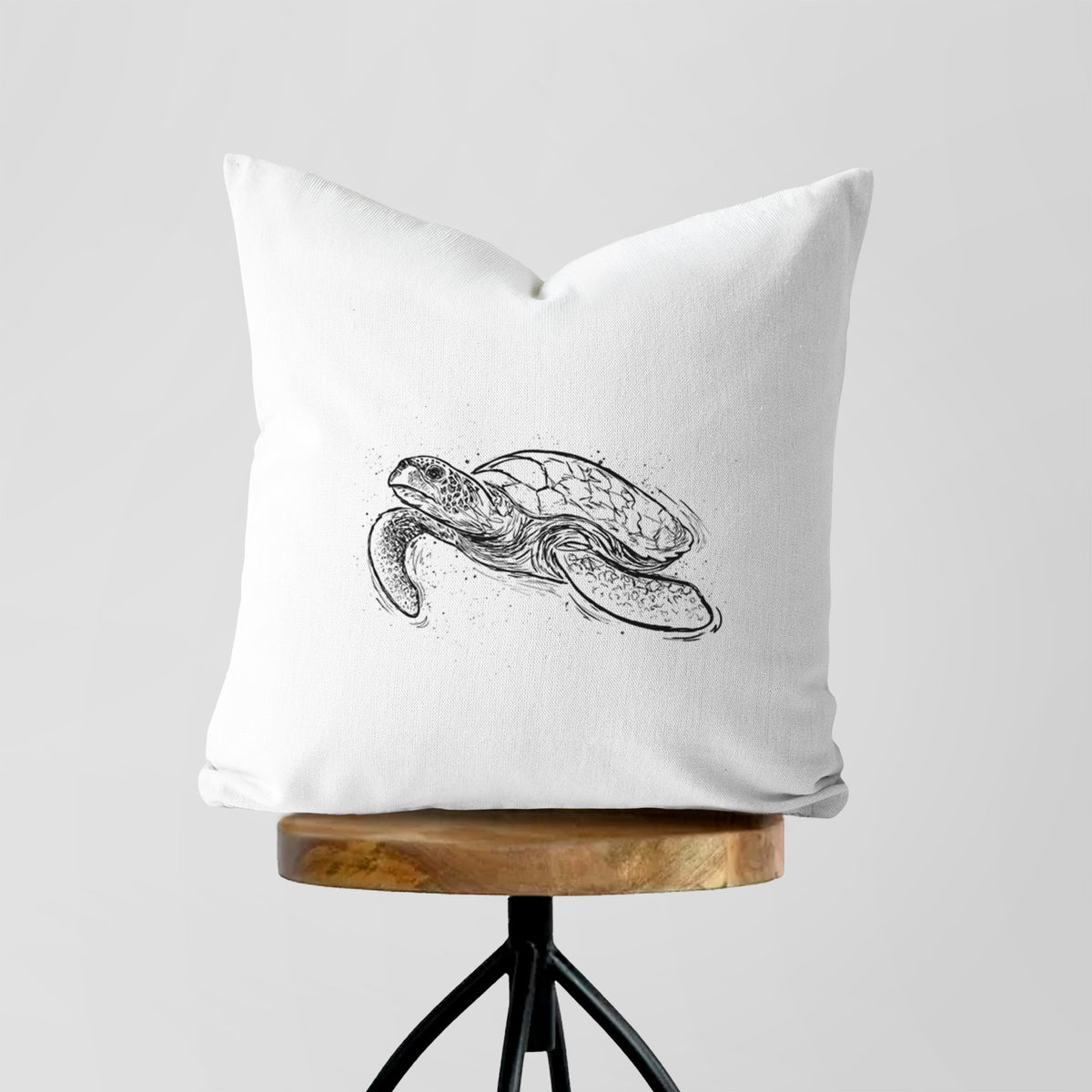 White pillow cover printed with a black ink illustration of a hawksbill sea turtle swimming, with a patterned shell, on a wooden stool.