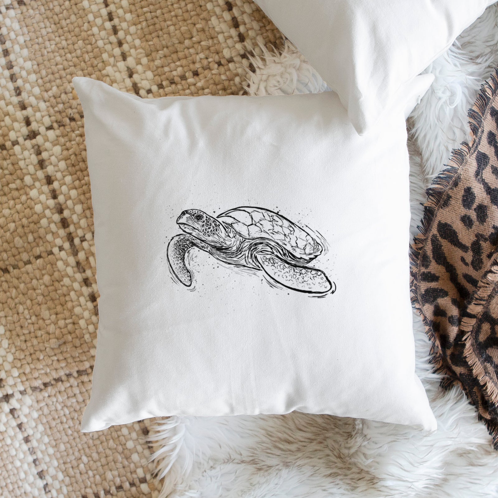 White pillow cover printed with a black ink illustration of a hawksbill sea turtle swimming, with a patterned shell, on a bench beside plants.