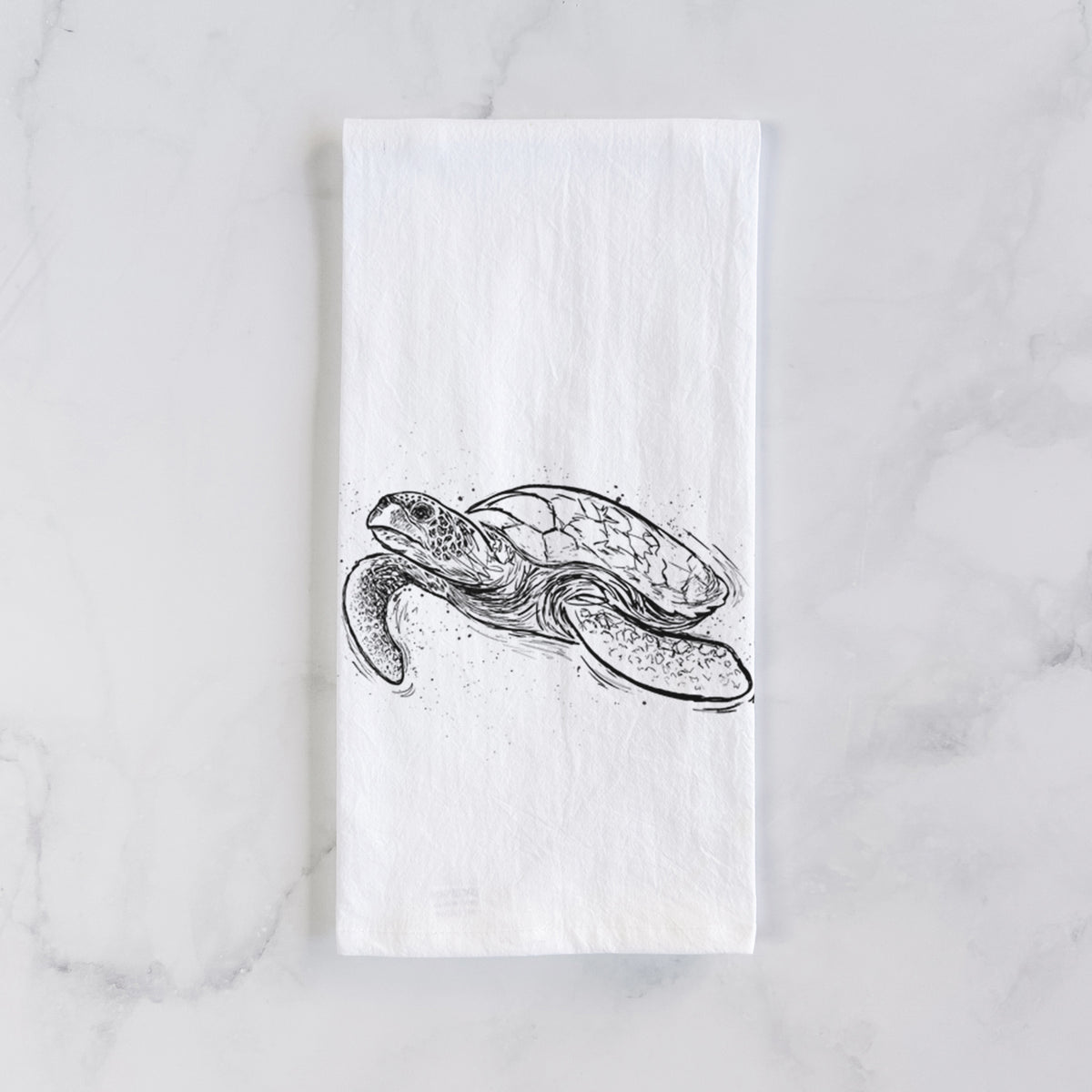 White cotton tea towel laid flat on marble, printed with a black ink illustration of a hawksbill sea turtle swimming, with a patterned shell.