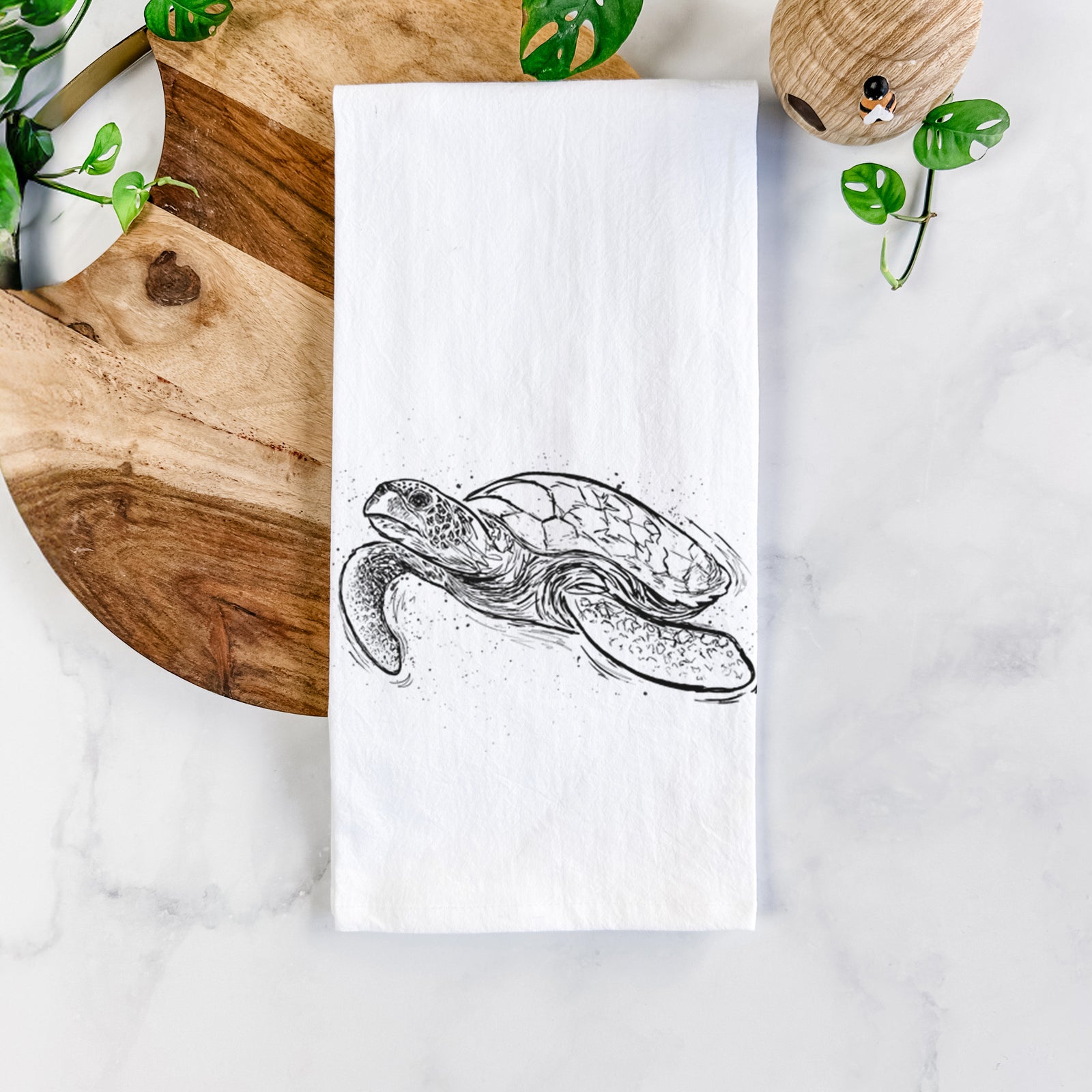 White cotton tea towel printed with a black ink illustration of a hawksbill sea turtle swimming, with a patterned shell, on a marble counter beside a round wooden cutting board and greenery.