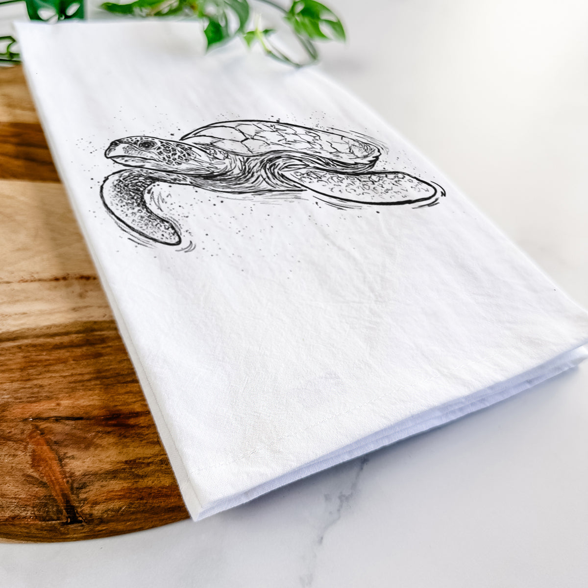 Close-up of a folded white tea towel on a wooden board, showing a black ink illustration of a hawksbill sea turtle swimming, with a patterned shell.