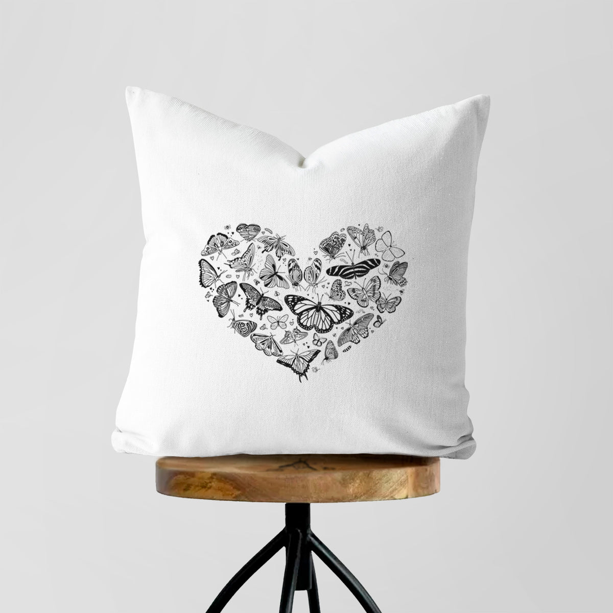 White pillow cover printed with a heart shape filled with small black ink butterflies and moths, on a wooden stool.