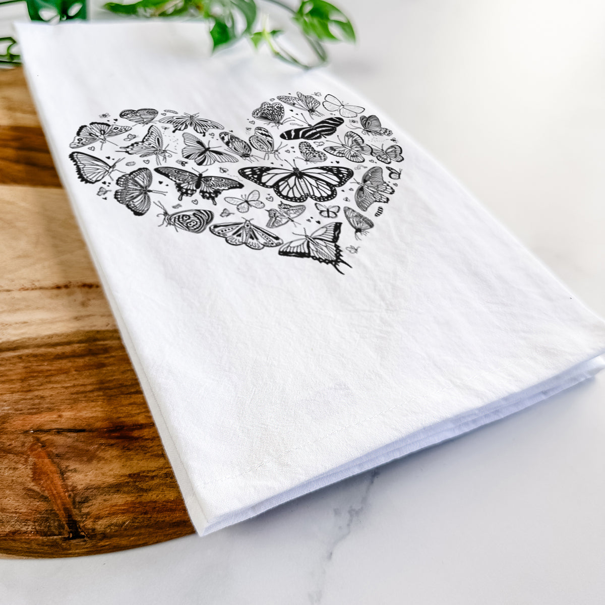 Close-up of a folded white tea towel on a wooden board, showing a heart shape filled with small black ink butterflies and moths.