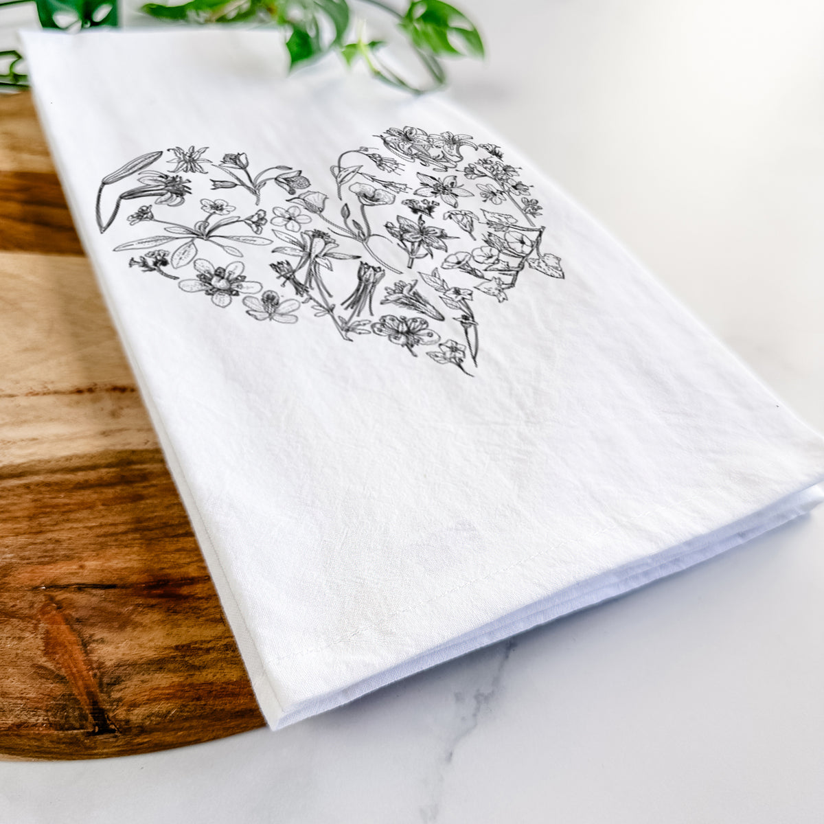 Close-up of a folded white tea towel on a wooden board, showing a heart shape filled with black ink California mountain wildflowers.