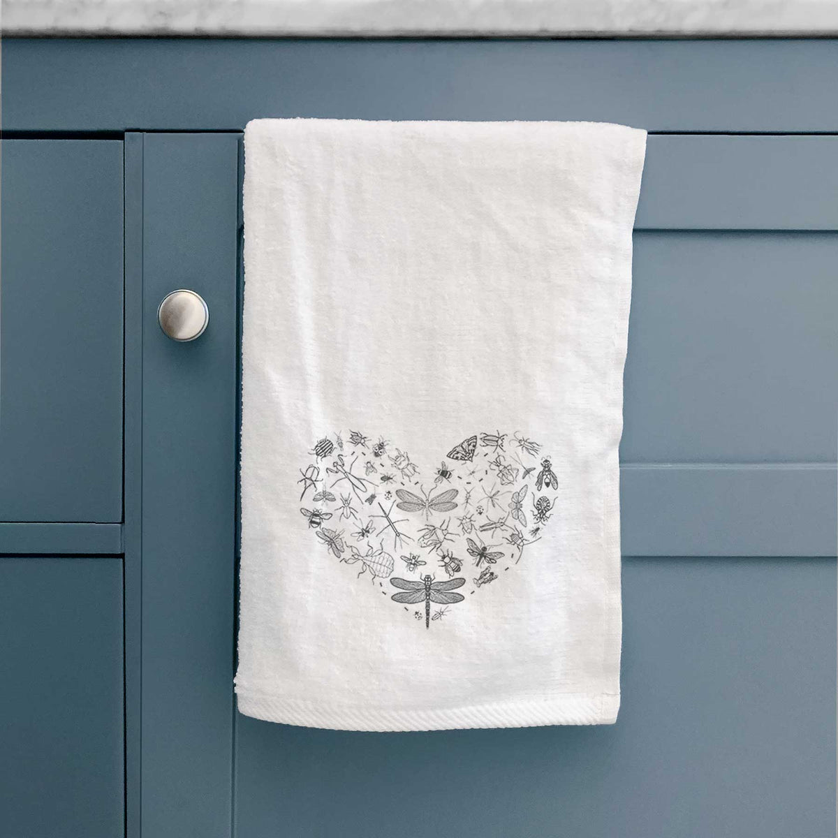 White hand towel printed with a heart shape filled with small black ink insects, including dragonflies, beetles and bees, hanging on a cabinet door.