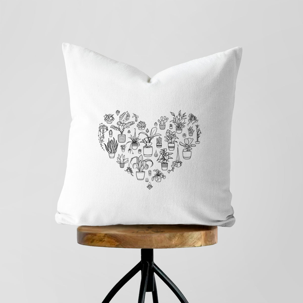 White pillow cover printed with a heart shape filled with black ink potted houseplants, on a wooden stool.