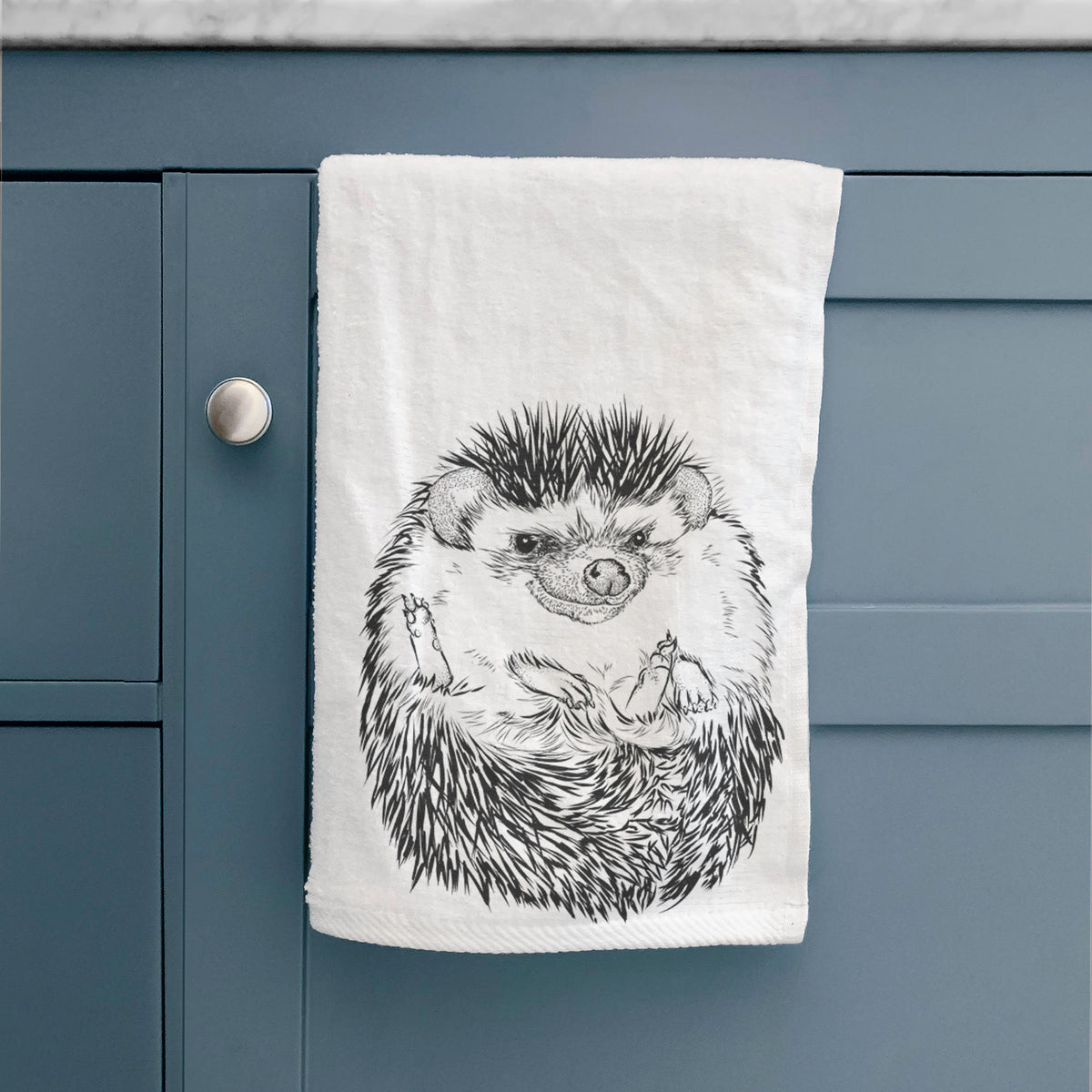 White hand towel printed with a detailed black ink illustration of a hedgehog curled up and facing forward, hanging on a cabinet door.