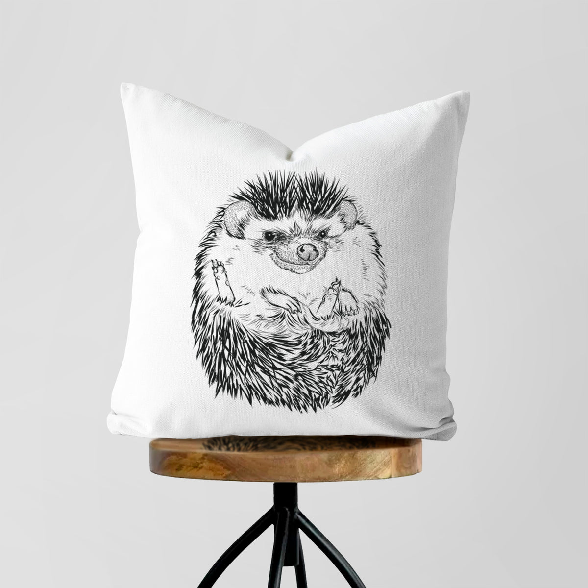 White pillow cover printed with a detailed black ink illustration of a hedgehog curled up and facing forward, on a wooden stool.