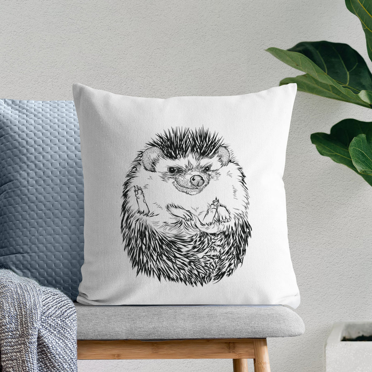 White pillow cover printed with a detailed black ink illustration of a hedgehog curled up and facing forward, on a bench beside plants.