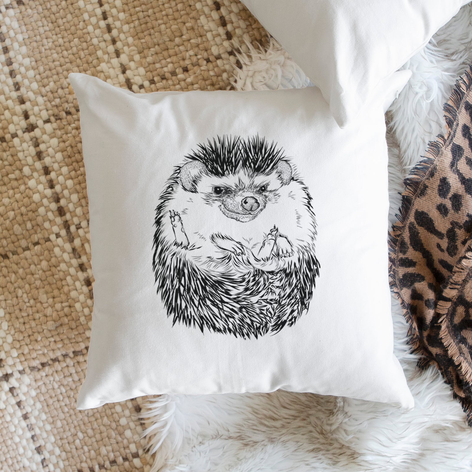 White pillow cover printed with a detailed black ink illustration of a hedgehog curled up and facing forward, on a bench beside plants.