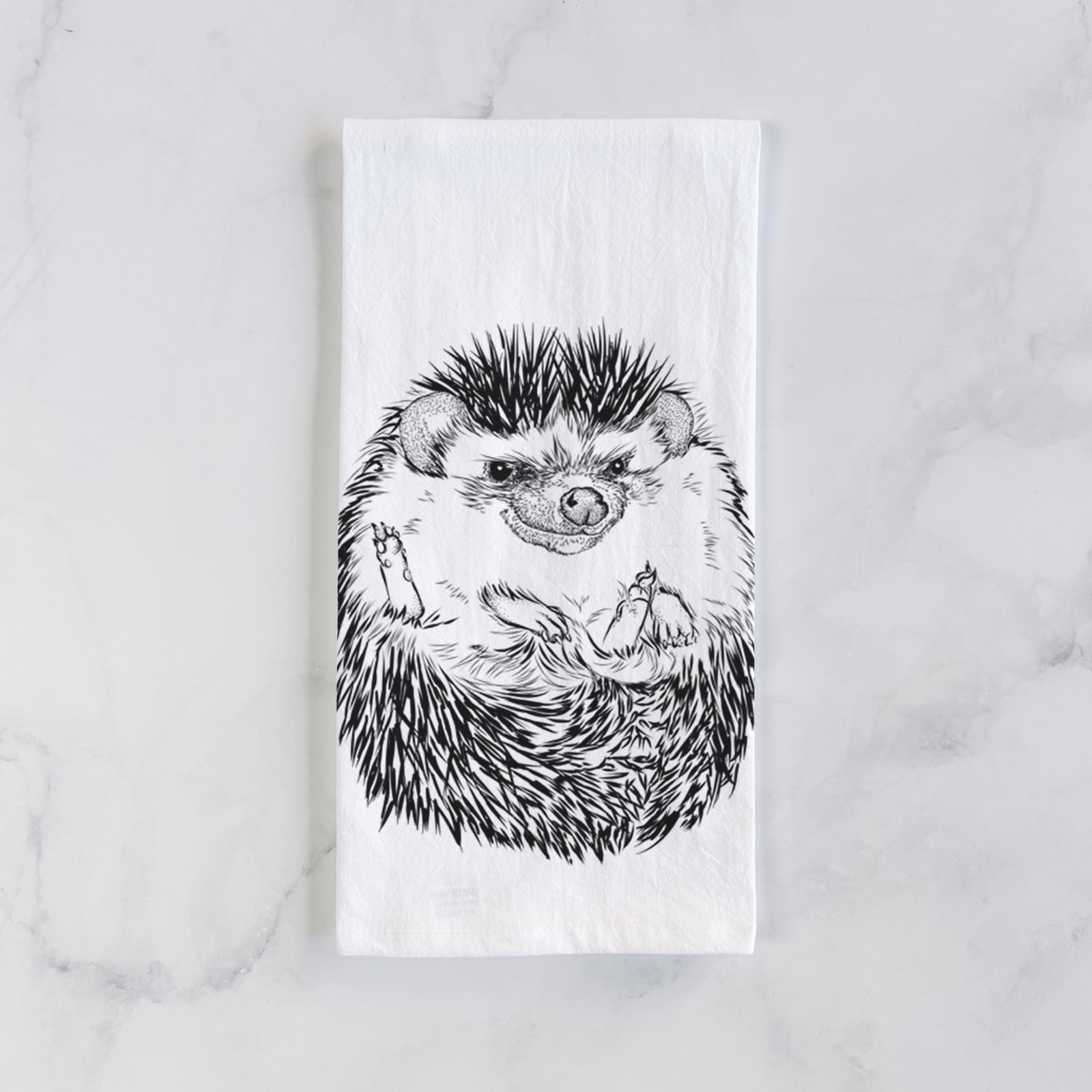 White cotton tea towel printed with a detailed black ink illustration of a hedgehog curled up and facing forward, on a marble counter beside a round wooden cutting board and greenery.