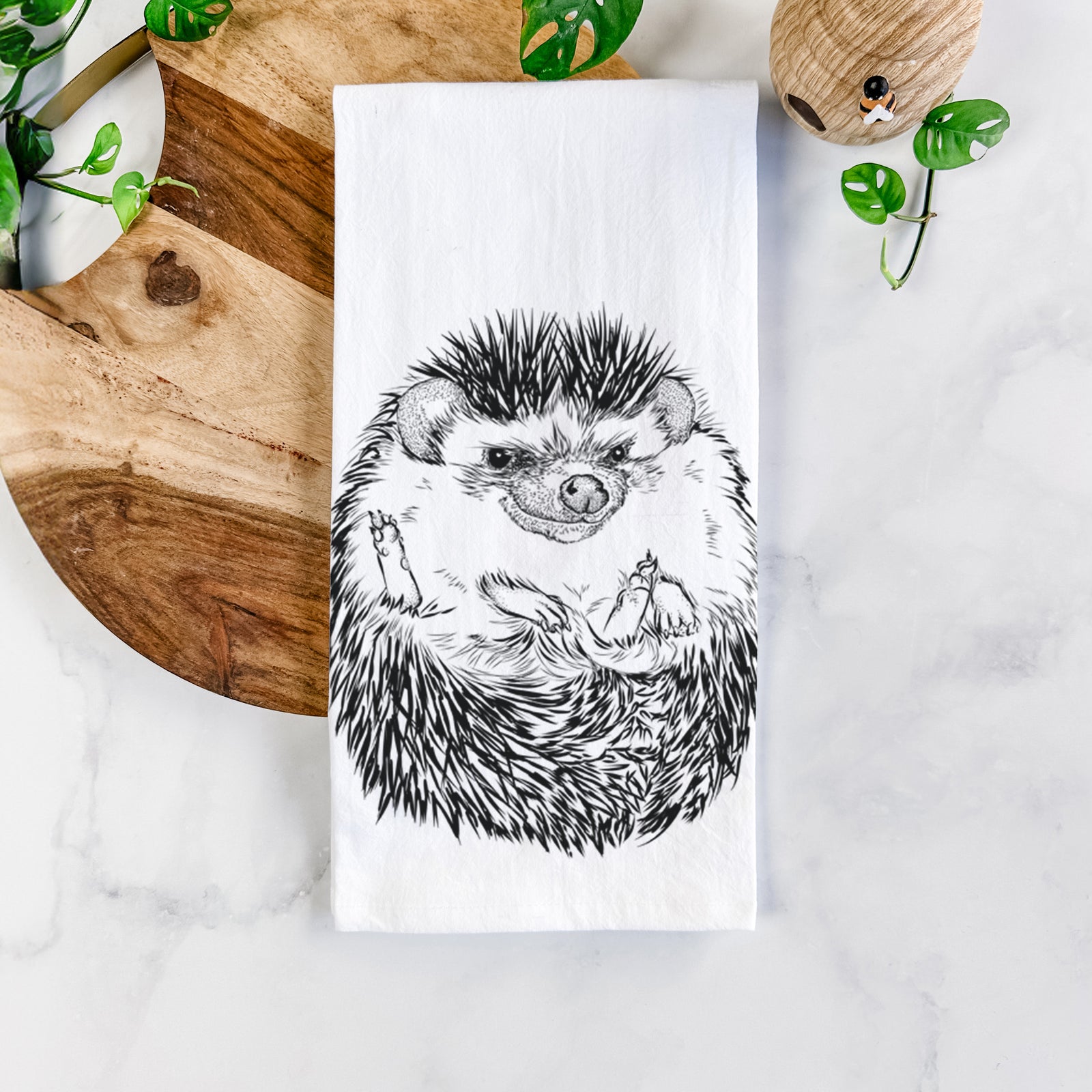 White cotton tea towel printed with a detailed black ink illustration of a hedgehog curled up and facing forward, on a marble counter beside a round wooden cutting board and greenery.