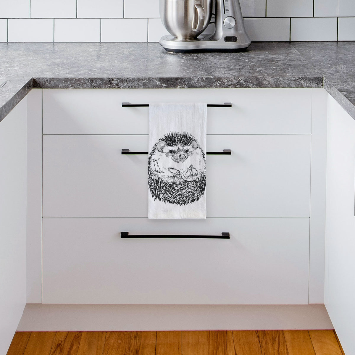 White tea towel printed with a detailed black ink illustration of a hedgehog curled up and facing forward, hanging from a drawer handle in a bright white kitchen.