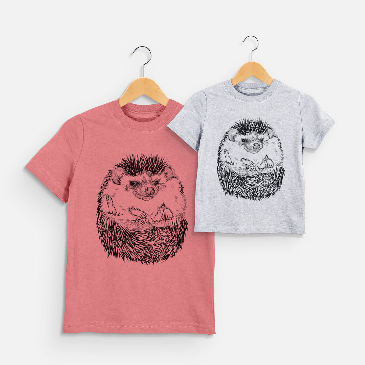 Two youth t-shirts in different colors on hangers, printed with a detailed black ink illustration of a hedgehog curled up and facing forward.