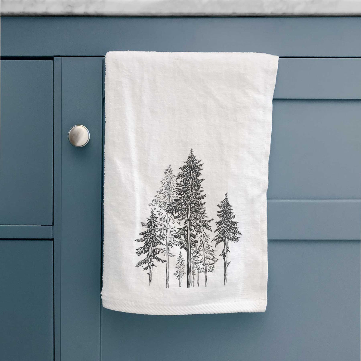 White hand towel printed with a black ink illustration of a forest of tall hemlock trees, hanging on a cabinet door.