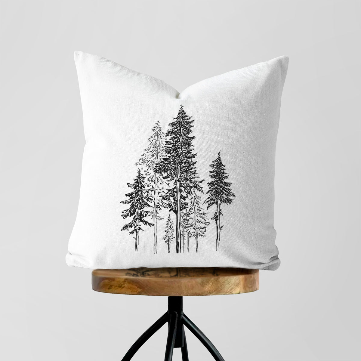 White pillow cover printed with a black ink illustration of a forest of tall hemlock trees, on a wooden stool.