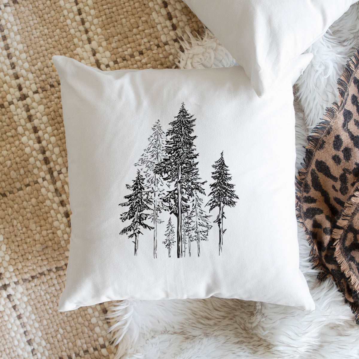 White pillow cover printed with a black ink illustration of a forest of tall hemlock trees, on a woven chair.