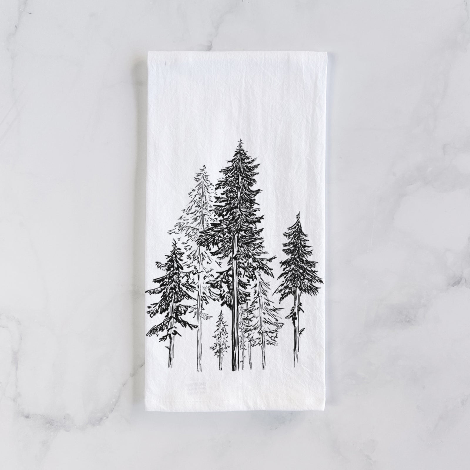 White cotton tea towel printed with a black ink illustration of a forest of tall hemlock trees, on a marble counter beside a round wooden cutting board and greenery.
