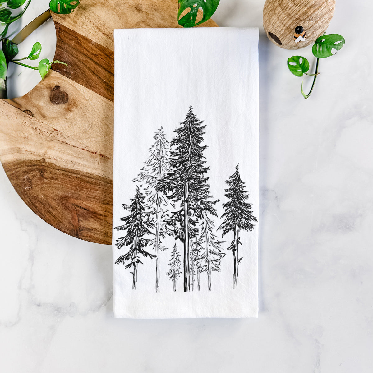 White cotton tea towel printed with a black ink illustration of a forest of tall hemlock trees, on a marble counter beside a round wooden cutting board and greenery.