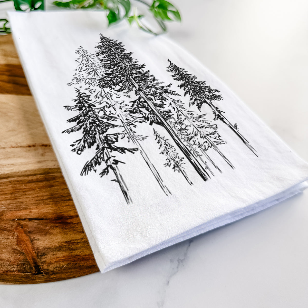 Close-up of a folded white tea towel on a wooden board, showing a black ink illustration of a forest of tall hemlock trees.