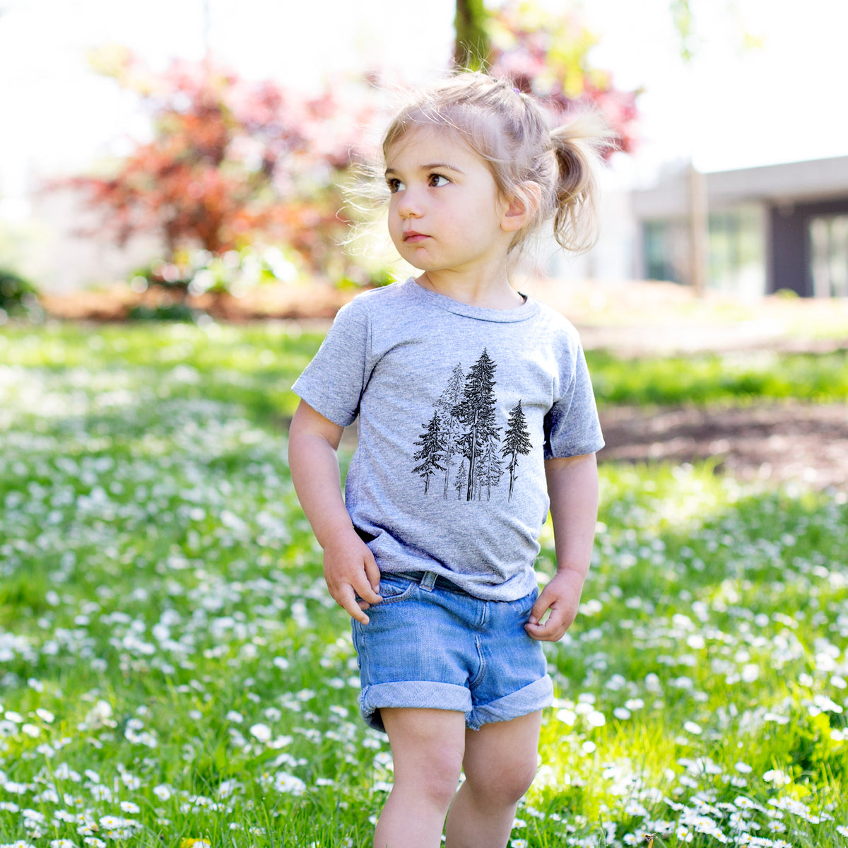 Toddler girl walking outdoors wearing a t-shirt printed with a black ink illustration of a forest of tall hemlock trees.