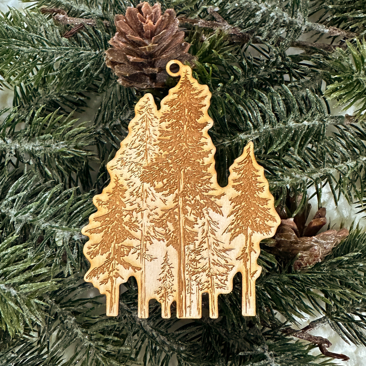 Wooden ornament featuring a detailed engraving of a forest of tall hemlock trees, on a knit blanket with pine cones.