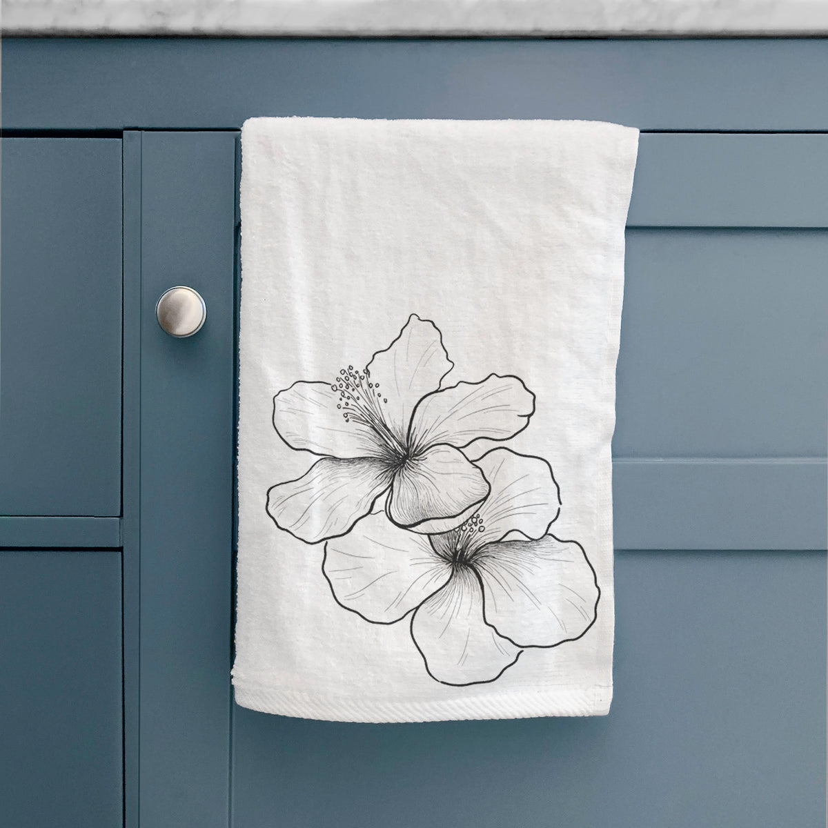 White hand towel printed with a black ink line drawing of three hibiscus flowers, hanging on a cabinet door.