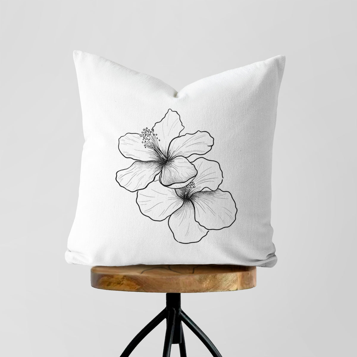 White pillow cover printed with a black ink line drawing of three hibiscus flowers, on a wooden stool.