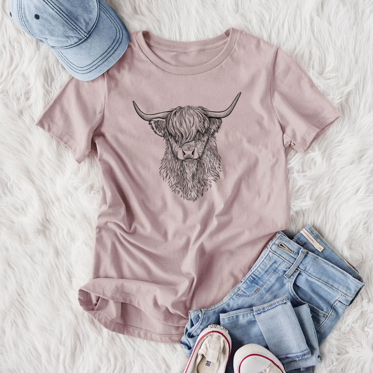 Women's relaxed fit cotton t-shirt printed with a detailed black ink portrait of a shaggy highland cow with long curved horns, styled flat on a white faux fur rug with jeans.