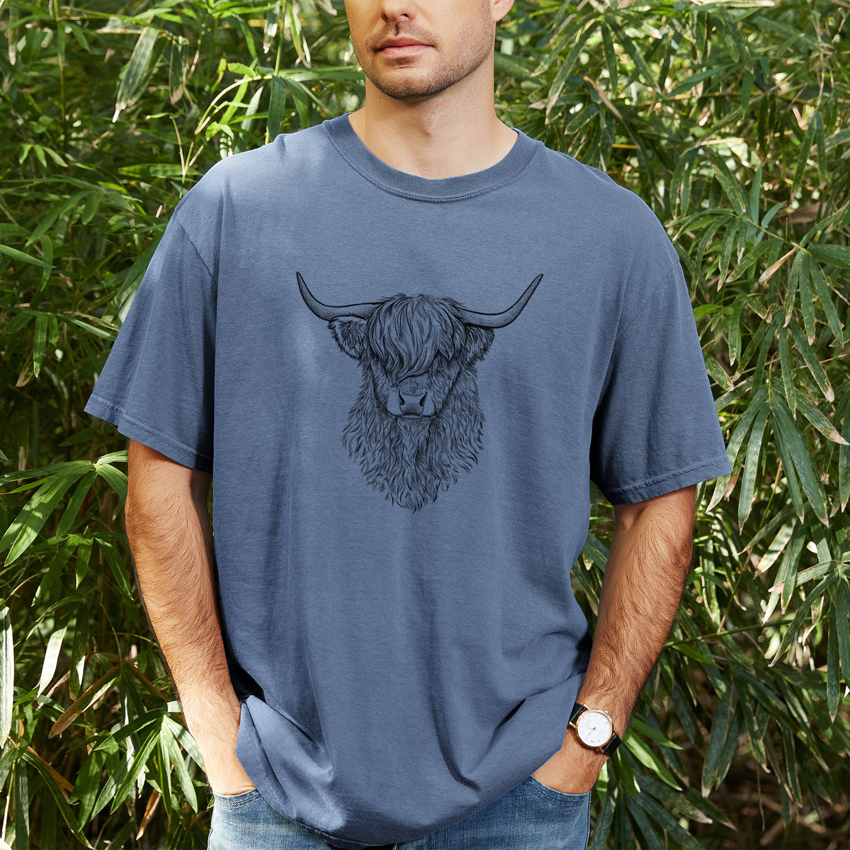 Man wearing a Comfort Colors t-shirt printed with a detailed black ink portrait of a shaggy highland cow with long curved horns, outdoors.