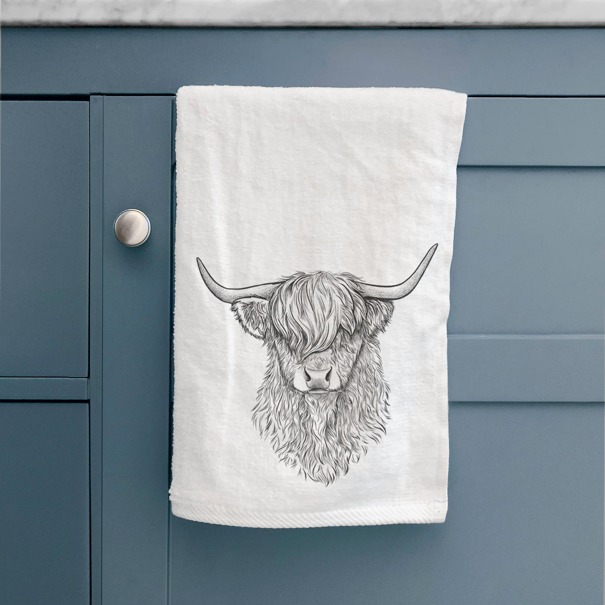 White hand towel printed with a detailed black ink portrait of a shaggy highland cow with long curved horns, hanging on a cabinet door.