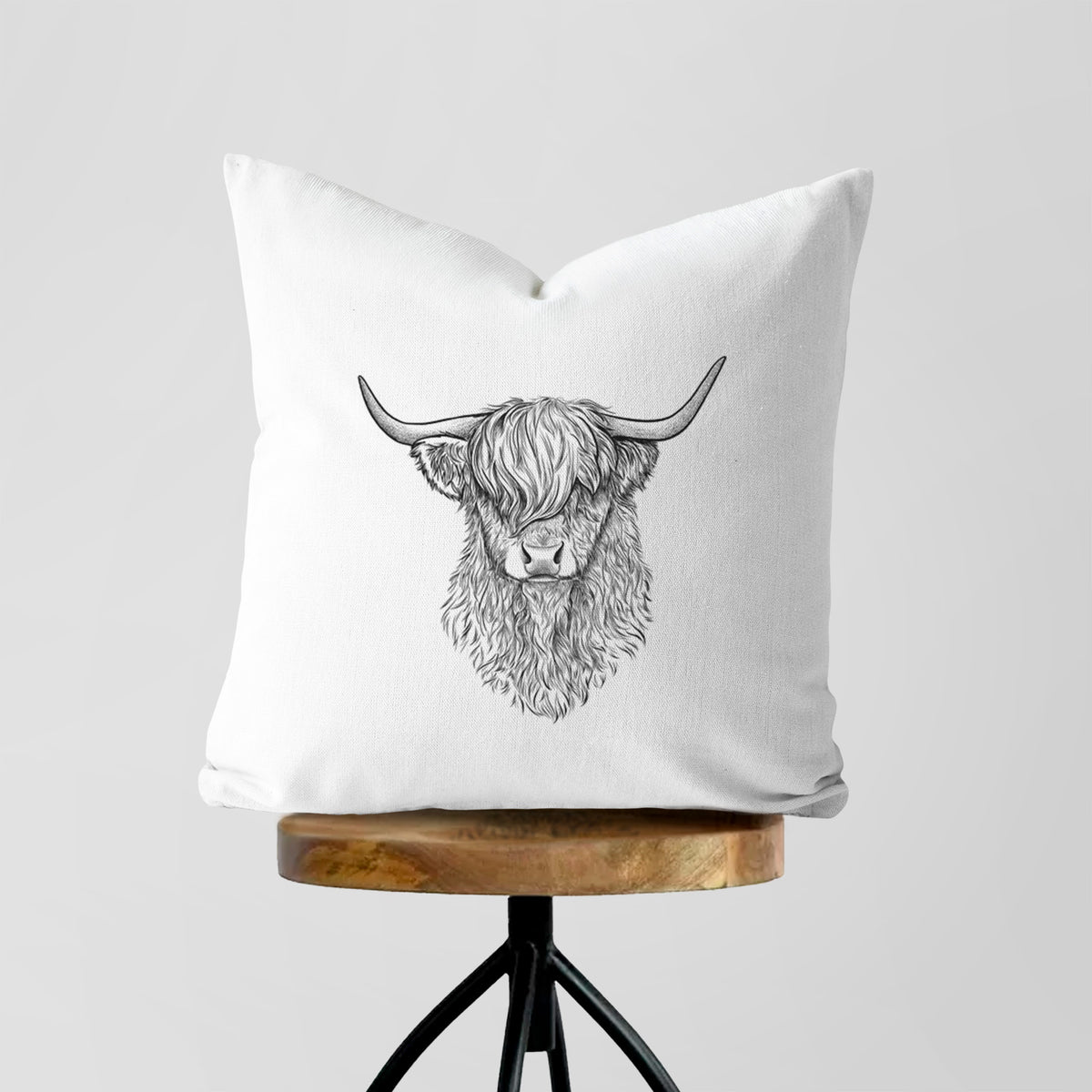 White pillow cover printed with a detailed black ink portrait of a shaggy highland cow with long curved horns, on a wooden stool.
