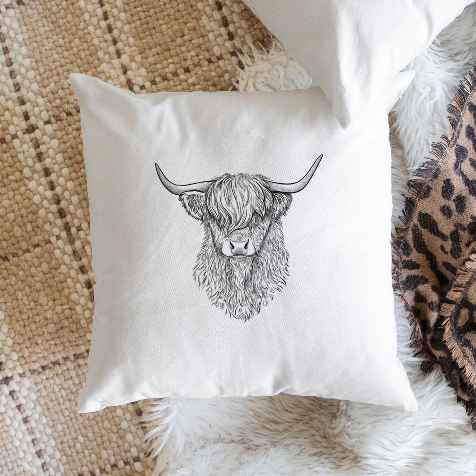 White pillow cover printed with a detailed black ink portrait of a shaggy highland cow with long curved horns, on a bench beside plants.