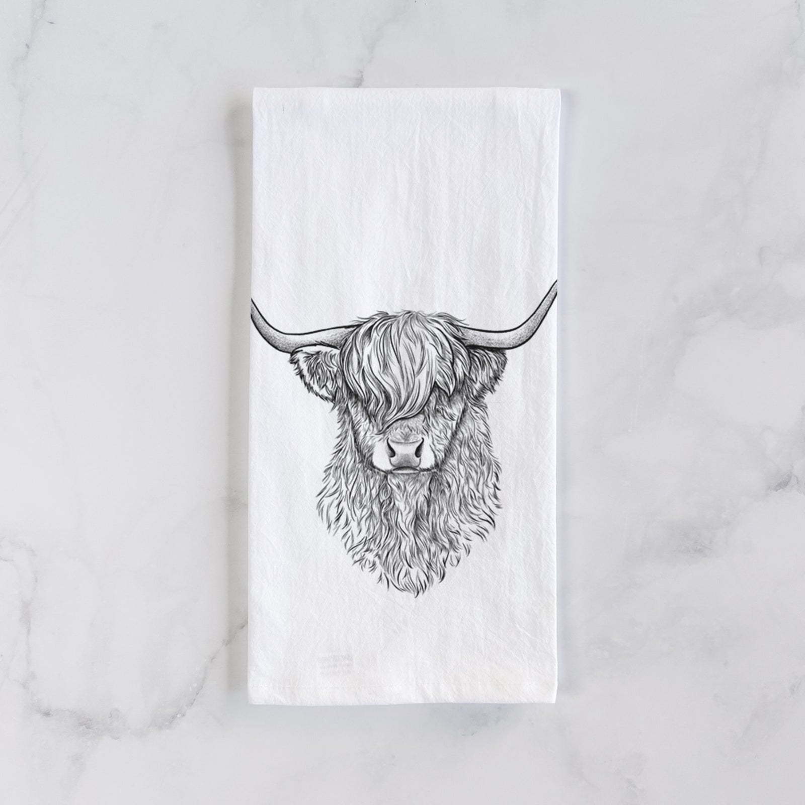 White cotton tea towel printed with a detailed black ink portrait of a shaggy highland cow with long curved horns, on a marble counter beside a round wooden cutting board and greenery.