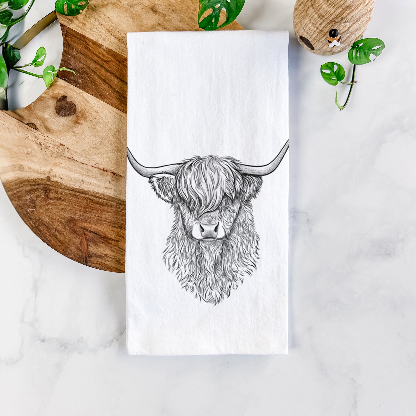 White cotton tea towel printed with a detailed black ink portrait of a shaggy highland cow with long curved horns, on a marble counter beside a round wooden cutting board and greenery.