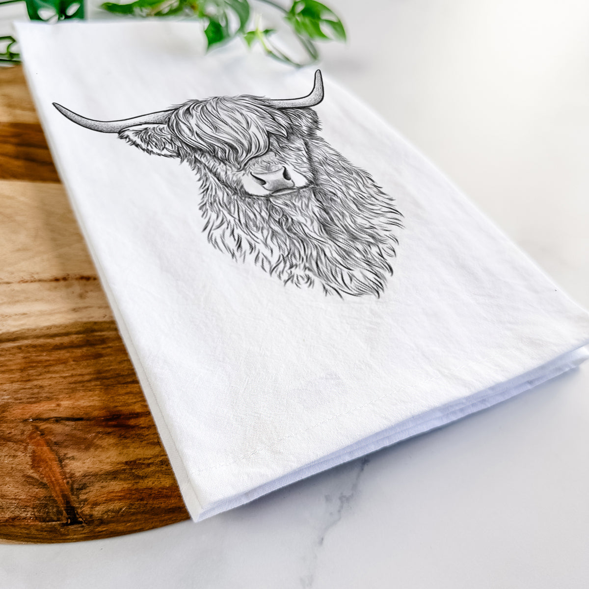 Close-up of a folded white tea towel on a wooden board, showing a detailed black ink portrait of a shaggy highland cow with long curved horns.