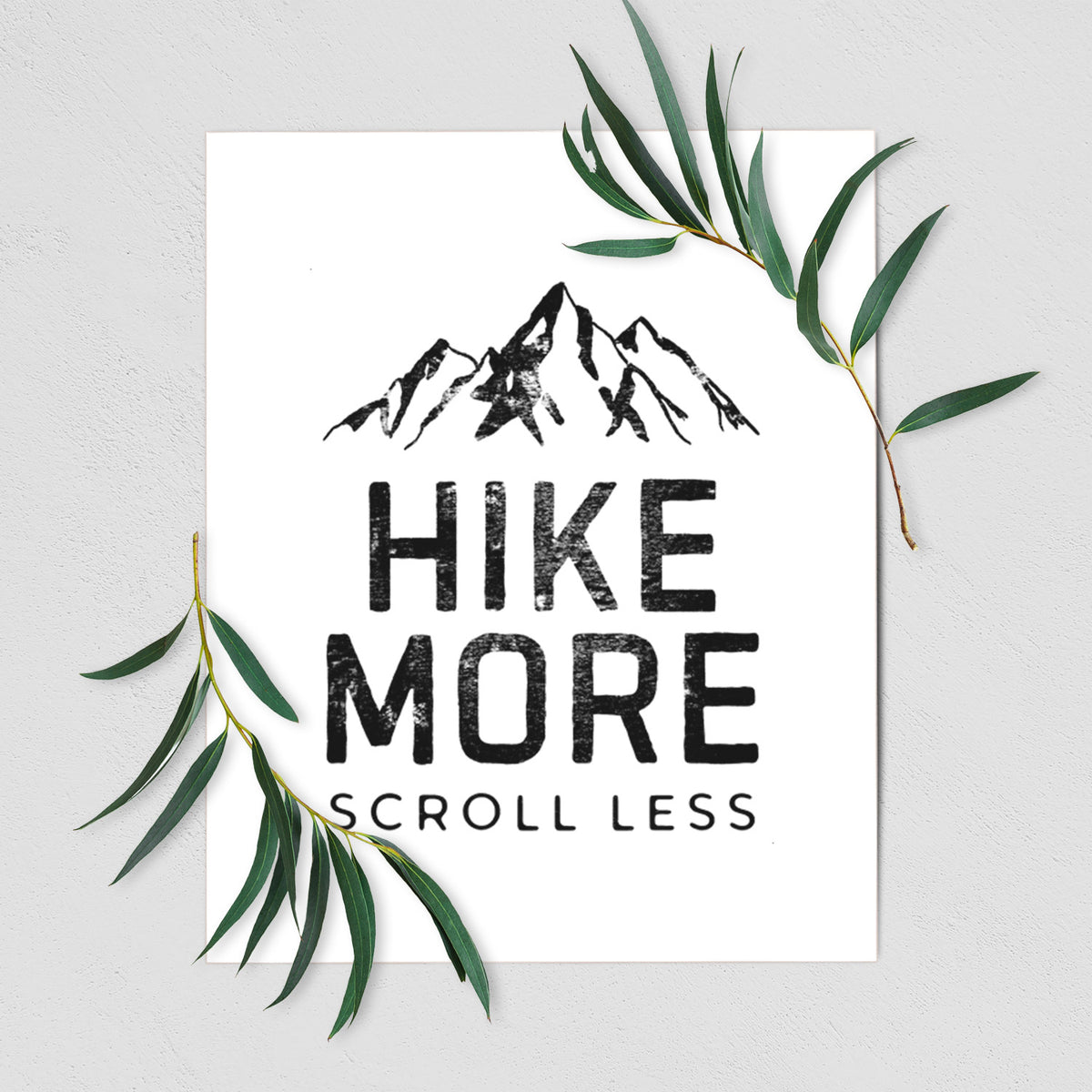 Art print of black mountain peaks above the bold words Hike More, Scroll Less, shown with eucalyptus leaves.