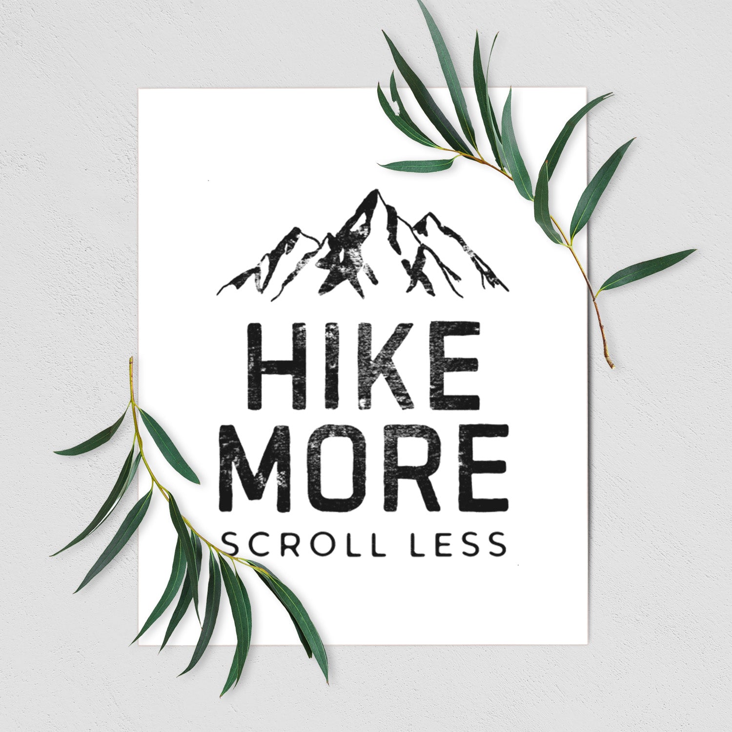 Art print of black mountain peaks above the bold words Hike More, Scroll Less, shown with eucalyptus leaves.