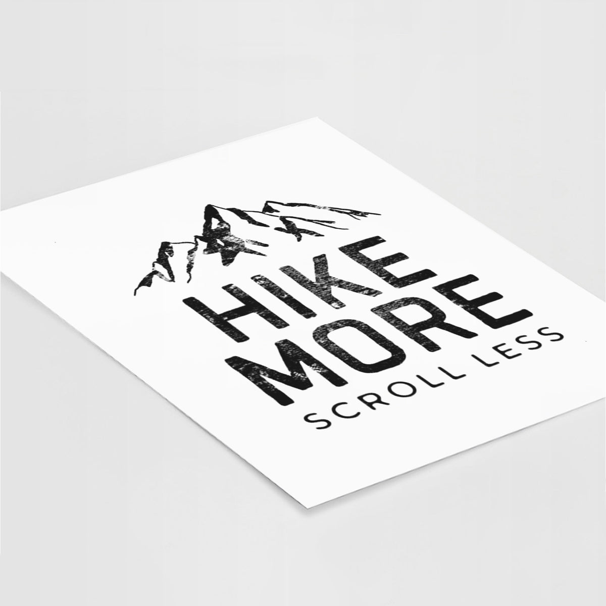 Art print of black mountain peaks above the bold words Hike More, Scroll Less, lying at an angle on a white surface.
