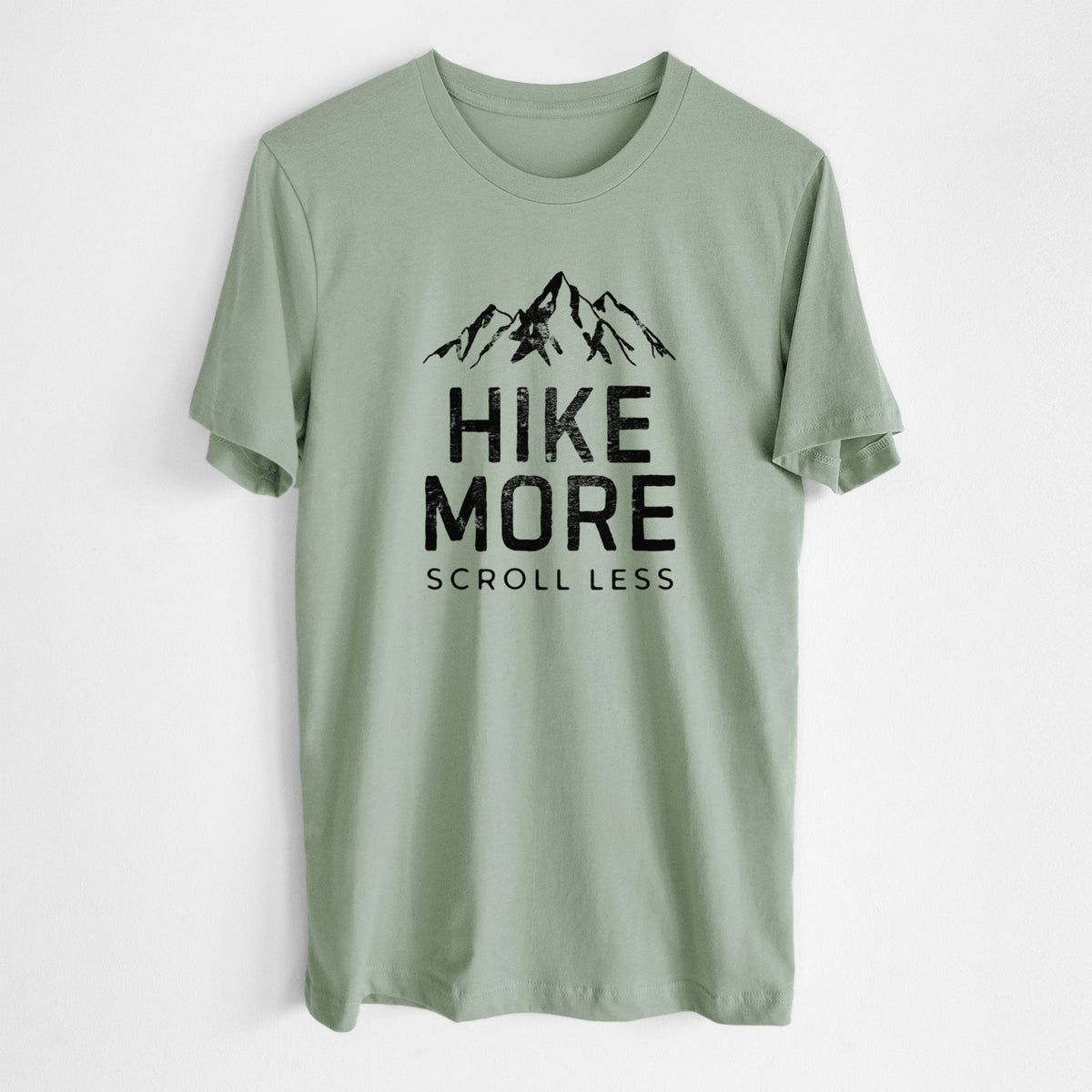 Sage green lightweight cotton t-shirt laid flat, printed with black mountain peaks above the bold words Hike More, Scroll Less.