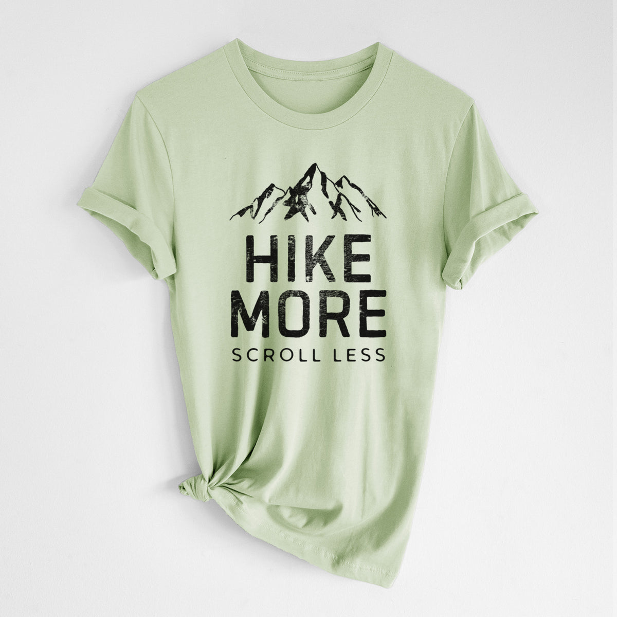 Spring green lightweight cotton t-shirt laid flat with the sleeves rolled, printed with black mountain peaks above the bold words Hike More, Scroll Less.
