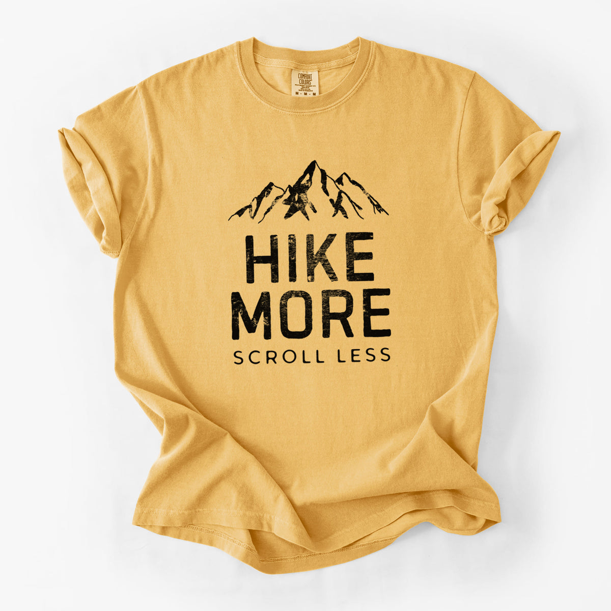 Golden wheat yellow Comfort Colors t-shirt laid flat, printed with black mountain peaks above the bold words Hike More, Scroll Less.