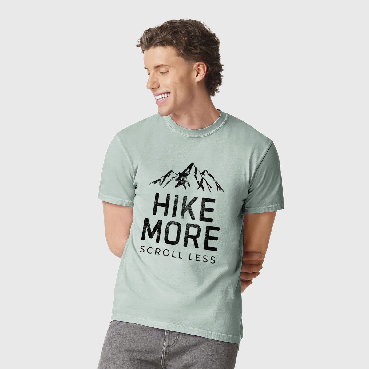 Man wearing a Comfort Colors t-shirt printed with black mountain peaks above the bold words Hike More, Scroll Less.