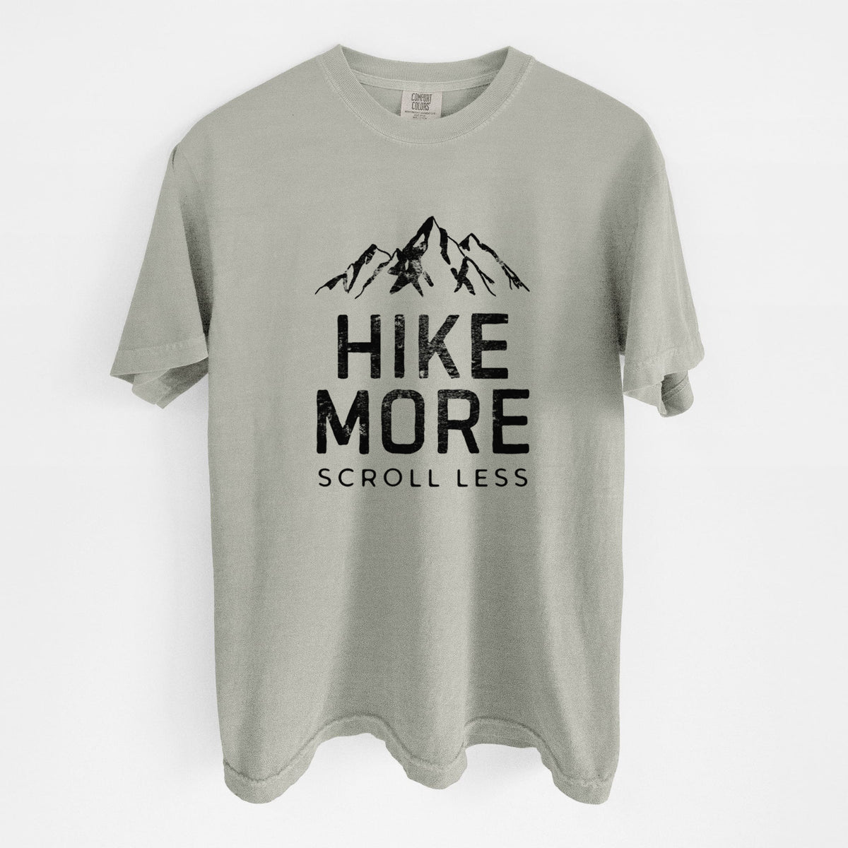 Sandstone beige Comfort Colors t-shirt laid flat, printed with black mountain peaks above the bold words Hike More, Scroll Less.